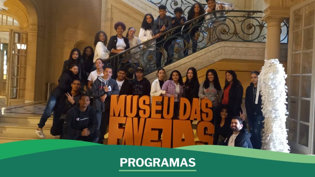 Foto dos jovens reunidos em frente ao letreiro do Museu das Favelas com arte escrito "programas".